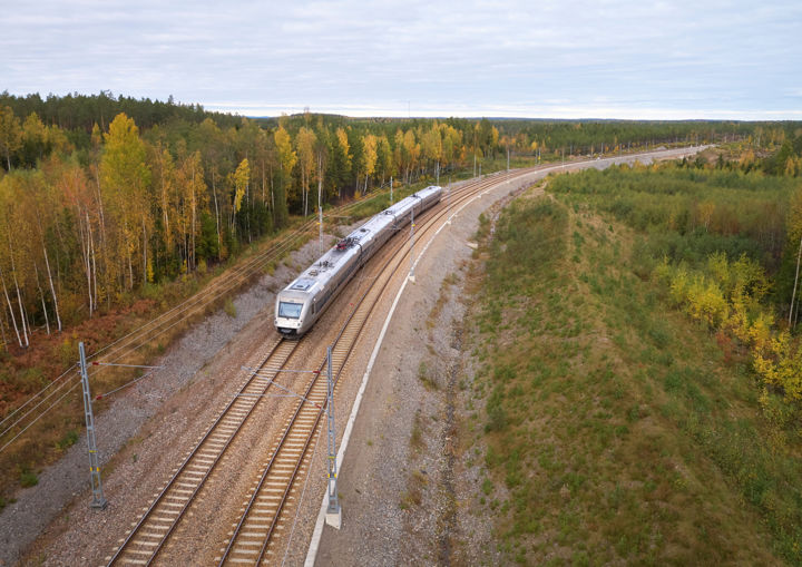Dubbelspår med persontåg genom skog med höstfärger.