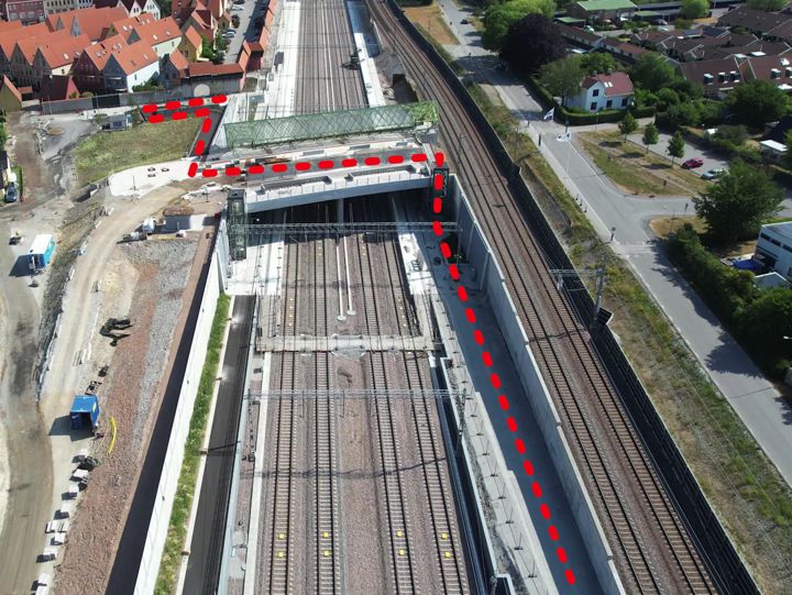 Flygbild över ett stationsområde med fyra spår i ett betongschakt. Över spåren går en bro i betong. En röd streckad linje visar hur gående och cyklister tar sig över bron och fram till hiss och trappa för att fortsätta på andra sidan.