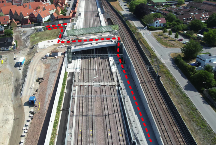 Flygbild över ett stationsområde med fyra spår i ett betongschakt. Över spåren går en bro i betong. En röd streckad linje visar hur gående och cyklister tar sig över bron och fram till hiss och trappa för att fortsätta på andra sidan.