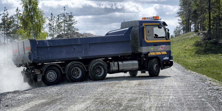 En lastbil med massor på flaket svänger upp på en grusväg med grönskande träd på varje sida om vägen.