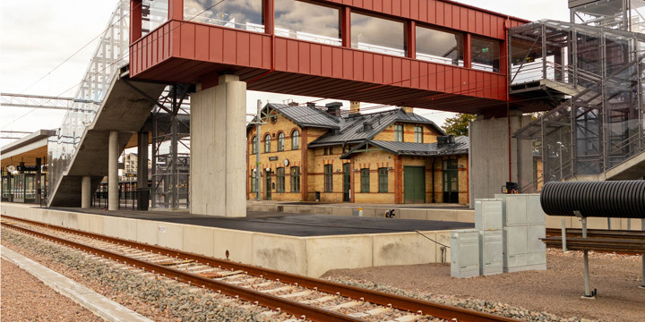 En tågstation med plattform. Över spåren finns en röd gångbro. I bakgrunden syns en gammal stationsbyggnad i gult tegel.