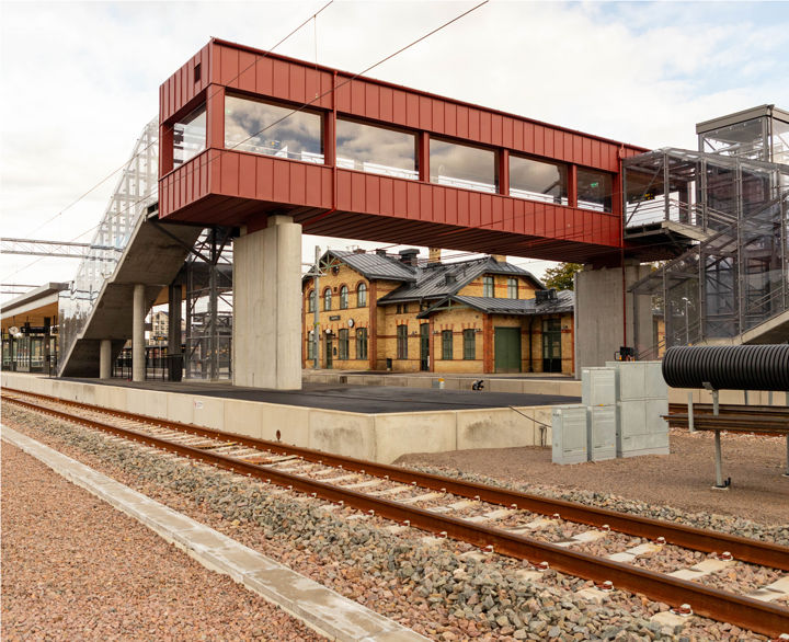 En tågstation med plattform. Över spåren finns en röd gångbro. I bakgrunden syns en gammal stationsbyggnad i gult tegel.