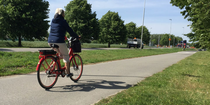 Äldre dam på röd cykel passerar från vänster till höger på cykelbana. Hon har en så kallad Hövding-hjälm på sig.