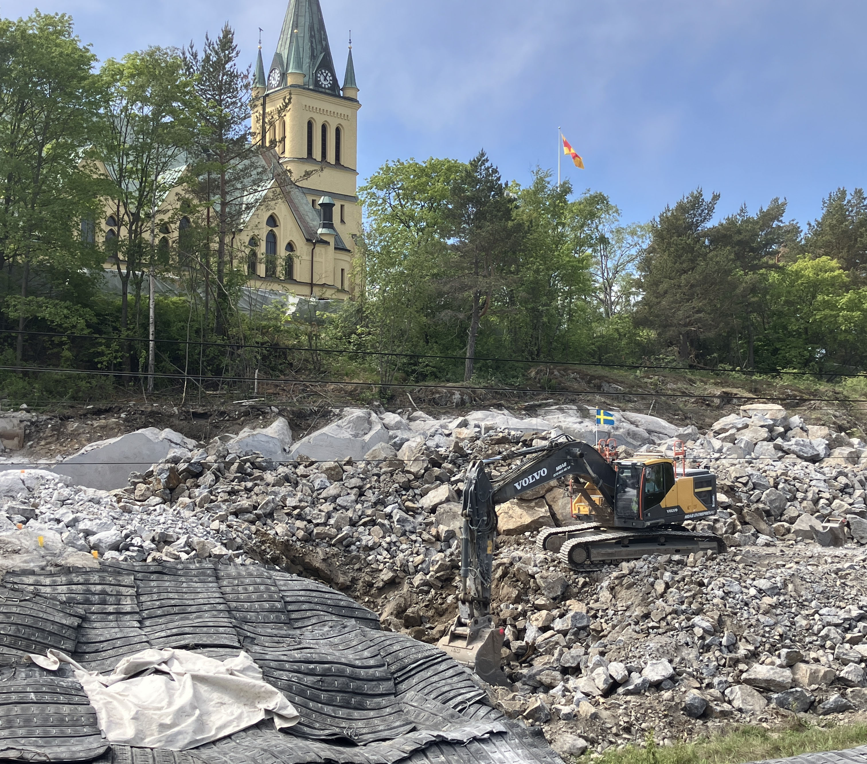 Pågående sprängarbeten nedanför Skönsmons kyrka. En grävmaskin står bland sprängsten och sprängmattor ligger utlagda.