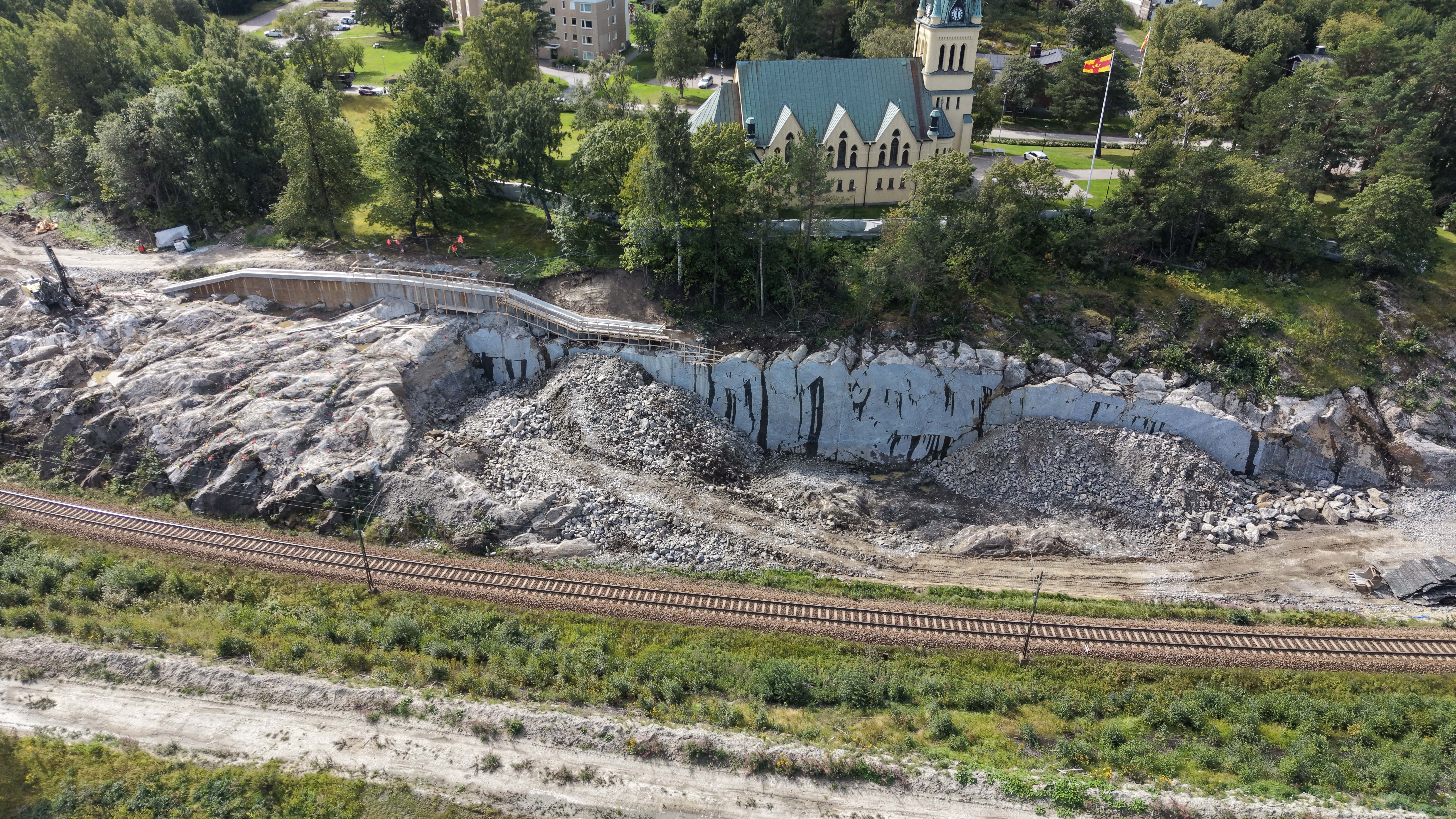 Pågående sprängarbeten nedanför Skönsmons kyrka. Krossat berg ligger framför en bergskärning, på skärningen träd och kyrkan. Delar av berget är fortfarande intakt. Befintligt spår framför berget.
