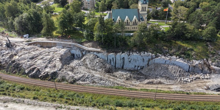 Pågående sprängarbeten nedanför Skönsmons kyrka. Krossat berg ligger framför en bergskärning, på skärningen träd och kyrkan. Delar av berget är fortfarande intakt. Befintligt spår framför berget.