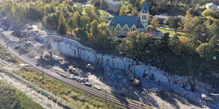 Krossat berg ligger framför en bergskärning där flera maskiner jobbar. Befintlig järnväg passerar skärningen och kyrkan som omges av träd uppe på bergskärningen.