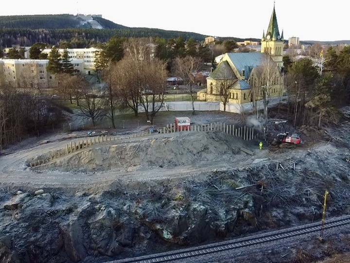 Skönsmons kyrka omgiven av träd och hyreshus. Nedanför kyrkan har träd avverkats och pålar sticker upp ur berget. En maskin borrar i marken och det ryker lite. En varselklädd arbetare står intill den. Järnvägen skymtar i förgrunden och Södra bergets slalombacke i bakgrunden.