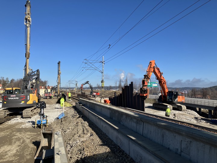 Ett flertal maskiner intill befintligt spår och arbetare längs järnvägen. Några maskiner slår ner spont mellan järnvägen och E4. Klarblå himmel och träd som ännu inte börjat grönska. Några fastigheter skymtar i bild samt rök från en fabrik.