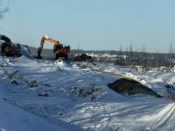 En fin vinterdag med blå himmel. Snön ligger vit på marken och två grävmaskiner syns i bild samt befintlig järnväg som skymtar nedanför ett berg. Oljecisternerna från oljehamnen skymtar i bakgrunden.
