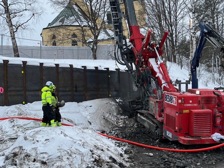 Två arbetare jobbar med en borrmaskin intill en stödmur av spont nedanför Skönsmons kyrka. Smutsig snö och mindre stenar intill borrmaskinen