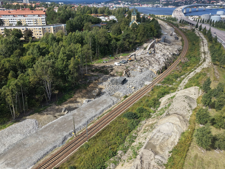 Vy över befintligt spår med bostäder och Skönsmons kyrka på ena sidan omgivna träd. Nedanför kyrkan syns det berg som vi spränger bort. På andra sidan spåret passerar E4 med Sundsvallsbron som skymtar längst bort i bilden.