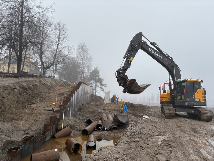 Skönsmons kyrka omgiven av några kala träd utan löv. Nedanför kyrkan har en stödmur monterats där berg som grävts fram delvis täcker plåten på stödmuren. Delar av stålrör ligger vid muren, delvis i lera och vatten. En grävmaskin jobbar intill stödmuren. Det är dimma.