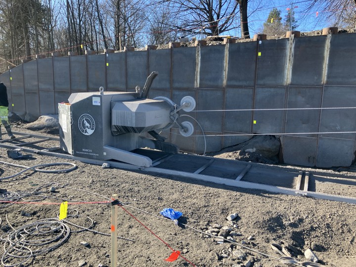 I bakgrunden en stödkonstruktion av stålspont. Framför den en står en maskin med inbyggd motor som driver två vajrar. Delar av en arbetare syns i bild.