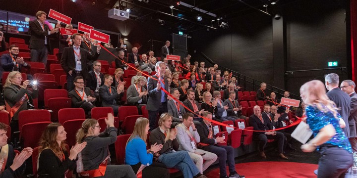 En mörk salong med människor som håller i röda skyltar. Längst fram vid scenen står två mörkklädda män som knyter ihop ett rött band.