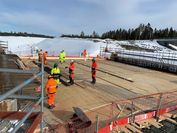 Flera varselklädda människor står på en byggarbetsplats, en vinterdag med snö i bakgrunden