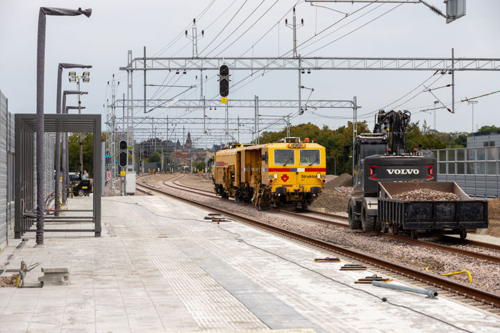 En nybyggd stationsplattform vid ett dubbelspår. Ett gult maskintåg och en spårgående grävmaskin står på ett av spåren.