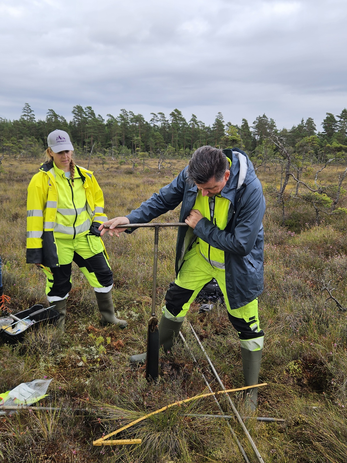 Två personer med varselkläder utför arkeologiska undersökningar ute på en moss.