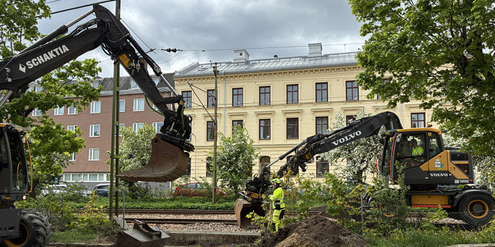 Två grävmaskiner och en järnvägsarbetare intill spåren genom Sundsvall. Framför ligger en jordhög och i bakgrunden större stenhus.