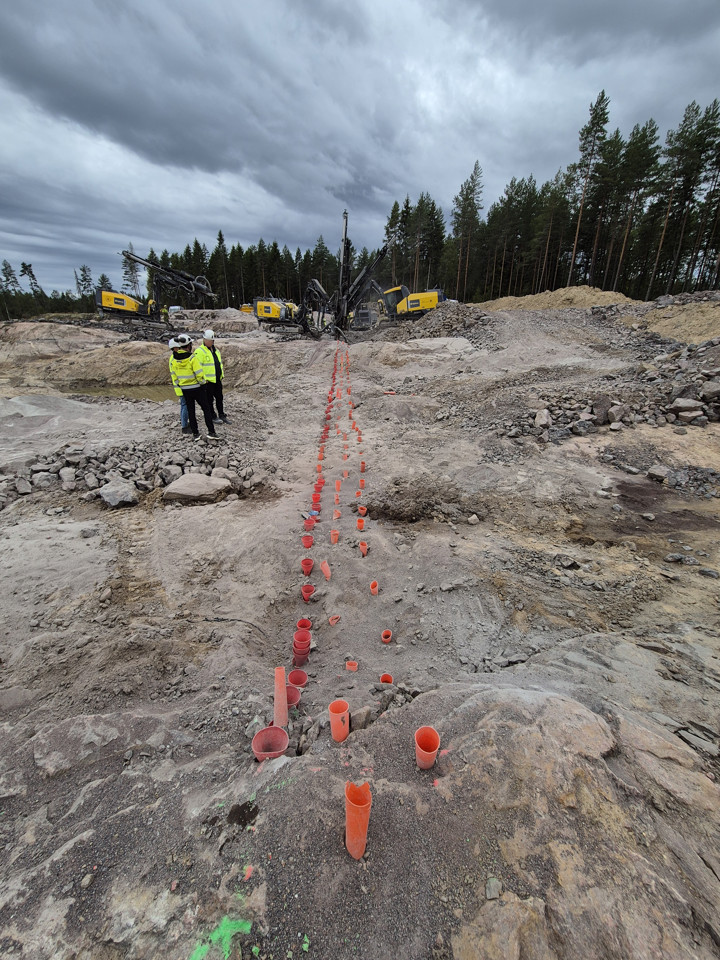 Tre personer står vid en linje med sprängladdningar, tre borriggar i bakgrunden.