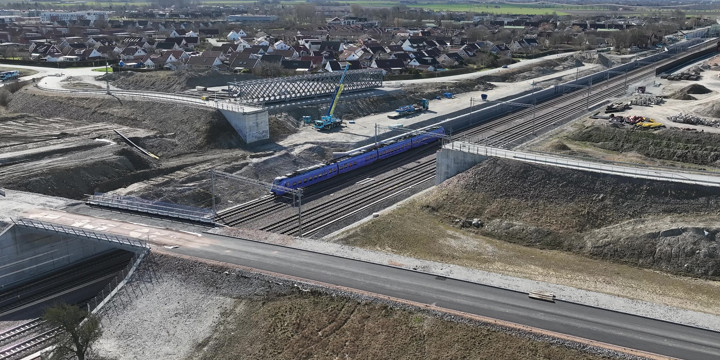 En fackverksbro i stål ligger vid sidan om järnvägen, och en betongbro som sträcker sig över en fyrspårig järnväg. Runt broarna är det en byggarbetsplats med grus och maskiner. Bortom arbetsplatsen syns ett villaområde.
