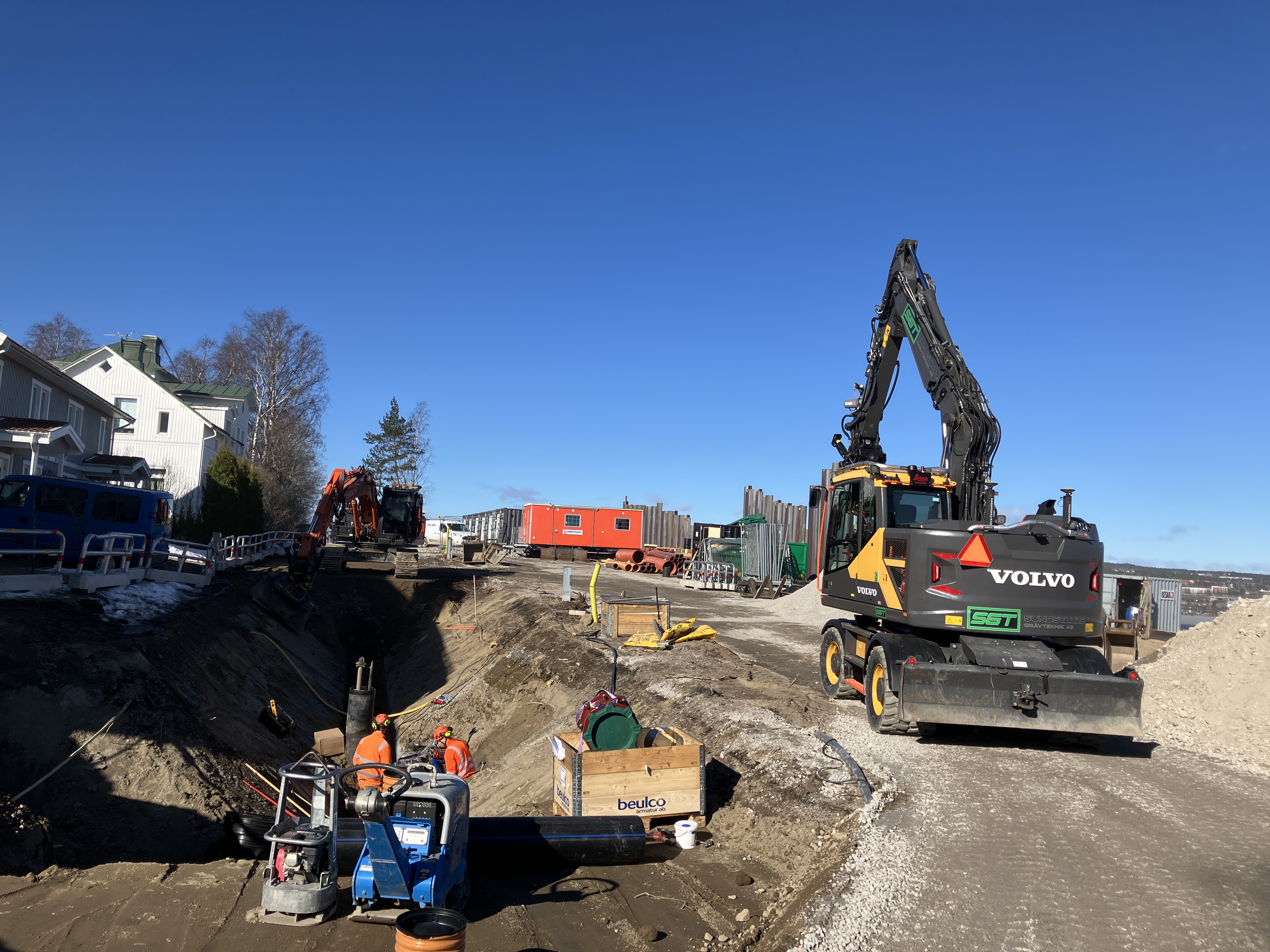 Uppgrävd väg där två arbetare står nere i en grop och jobbar. Två fastigheter nära arbetsområdet där grävmaskiner, en barack och arbetsmaterial samsas om utrymmet. Stålspont som utgör en stödmur syns i bild.