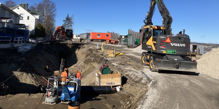Uppgrävd väg där två arbetare står nere i en grop och jobbar. Två fastigheter nära arbetsområdet där grävmaskiner, en barack och arbetsmaterial samsas om utrymmet. Stålspont som utgör en stödmur syns i bild.