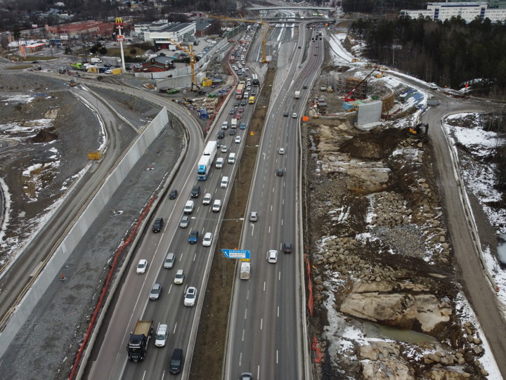Bilden visar väg E4/E20 i Norrgående riktning. På vägen åker många bilar. På sidan om vägen på båda sidor pågår bygget av den nya trafikplatsen, nya vägar och anslutningar till nya E4 Förbifart Stockholm. Bilden är tagen med en drönare som flyger över Vårby backe mot Kungens kurva.