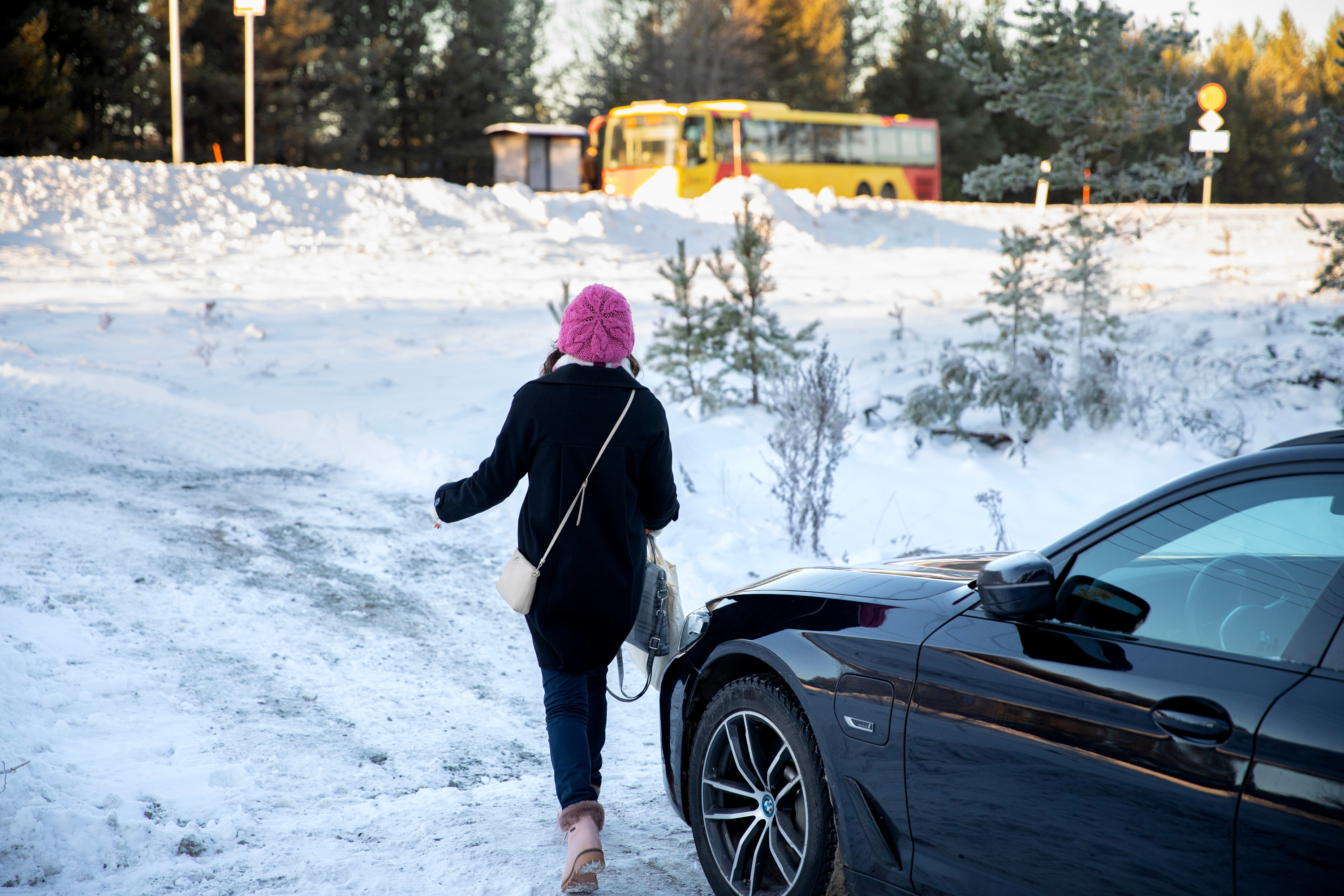 En kvinna har parkerat sin bil och går till en väntande buss vid en busskur. Det ligger snö på marken.