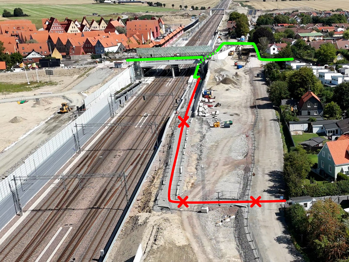 Flygbild över en järnvägsstation. Till vänster i bild fyra spår med sidoplattformar. Järnvägen ligger i ett schakt. Över järnvägen går en bro. Till höger i bild ett byggområde och med grushögar och mindre schakt. I bildens högerkant syns också ett villakvarter. Grön och röd markering visar vägar för gående och cyklister.