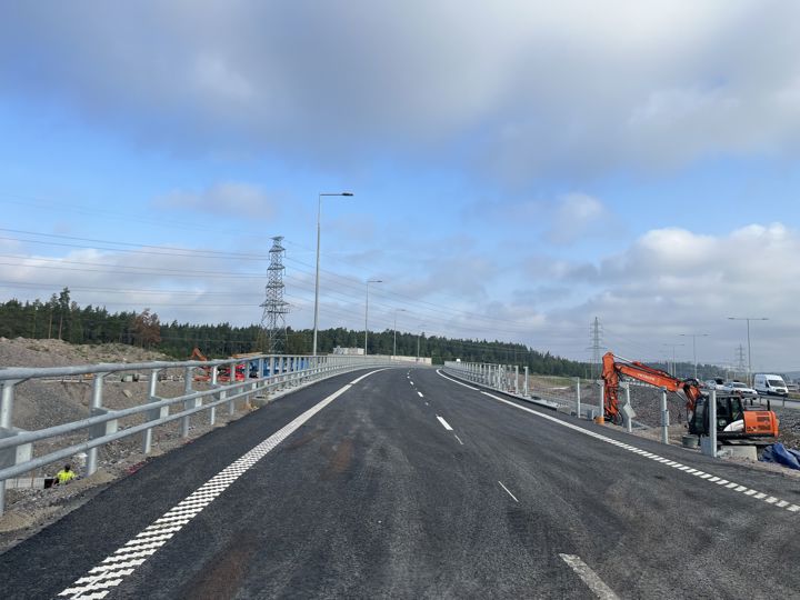Bron som leder trafiken från Norrortsleden i södergående riktning in mot Stockholm är färdig och öppnar för trafik. Bilden visar en asfalterad väg. Vägen är försedd med vägräcken. Till höger i bild arbetar en grävmaskin.