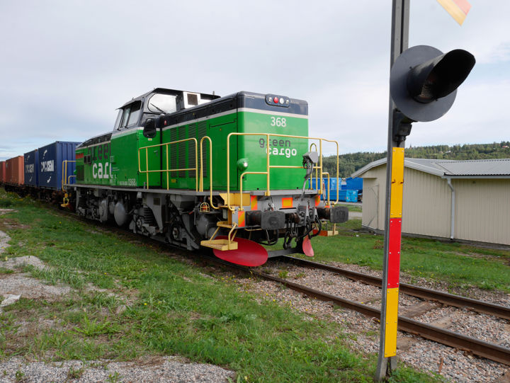 Green Cargo godståg passerar på ett industrispår. Loket närmar sig en signalstolpe, en vit byggnad i bakgrunden.