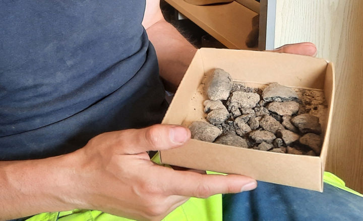 Arkeologen håller en liten låda stor som en tallrik i sina händer, i lådan ligger små grå bitar som liknar sandsten