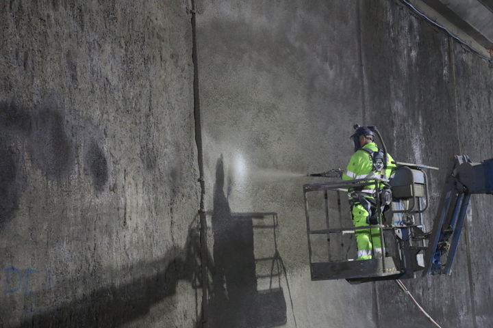 En man i varselkläder står upphöjd på en kran. Sprutar en vätska mot en grå betongvägg.