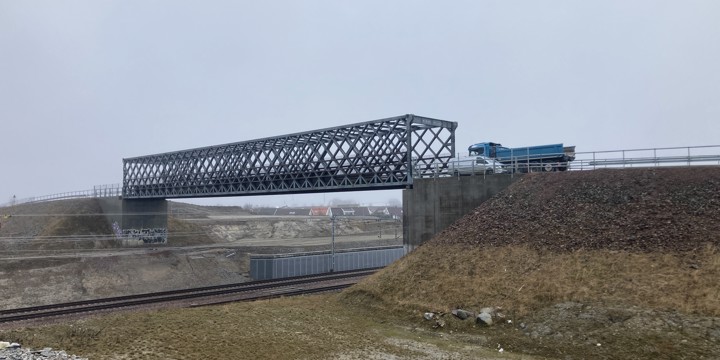 En fackverksbro i stål som sträcker sig över en fyrspårig järnväg. På bron passerar en blå lastbil och en vit personbil.