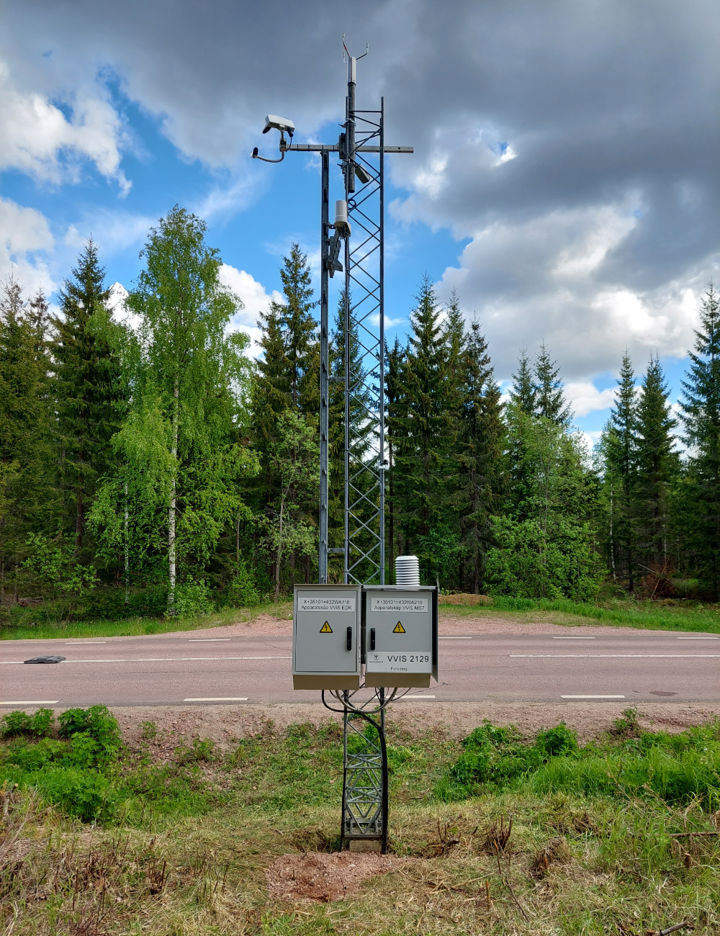 En mast med väderstation vid en väg med skog bakom