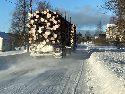 Timmerlastbil på smal vinterväg genom ett mindre samhälle