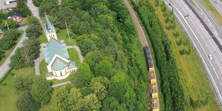 Skönsmons kyrka omgiven av gröna träd. På järnvägen nedanför kyrkan passerar ett godståg och E4 skymtar parallellt med järnvägsspåret.