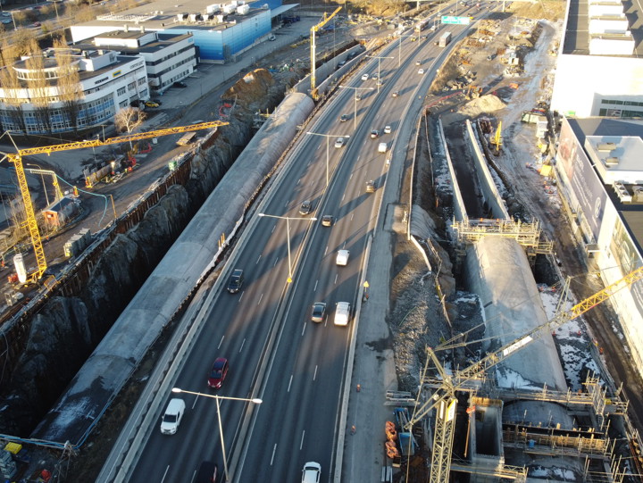 Bilden visar väg E4/E20 i norrgående riktning. På varje sida om vägen pågår arbeten med betongtunnlarna som ska gå till och från E4 Förbifart Stockholm. En lyftkran lyfter byggmaterial på plats. Solen lyser och bilar kör på vägen som går i mitten av allt pågående vägarbete.