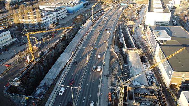Bilden visar väg E4/E20 i norrgående riktning. På varje sida om vägen pågår arbeten med betongtunnlarna som ska gå till och från E4 Förbifart Stockholm. En lyftkran lyfter byggmaterial på plats. Solen lyser och bilar kör på vägen som går i mitten av allt pågående vägarbete.