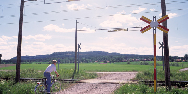 En cyklist ska till att passera en plankorsning. Det är grusväg genom plankorsningen och åkrar runt den. En solig sommardag med klar himmel.