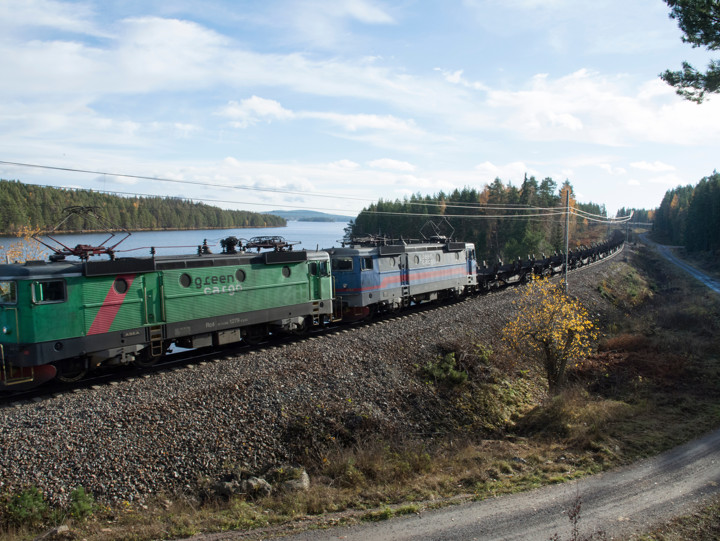 Ett godståg passerar på järnvägsbank med vattendrag i bakgrunden och en grusväg i förgrunden, nedanför järnvägsbanken.