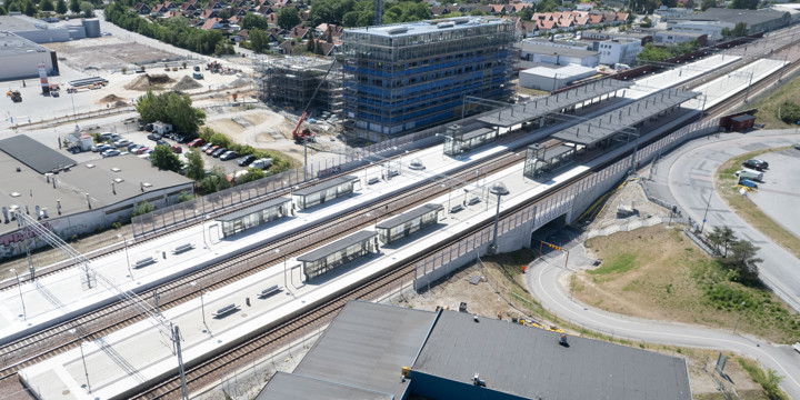 Stationsområde med två breda plattformar och fyra spår. Ovanför stationen byggs ett högt hus och där finns också en stor parkeringsplats. Framför stationen finns en väg och en verkstadslokal.