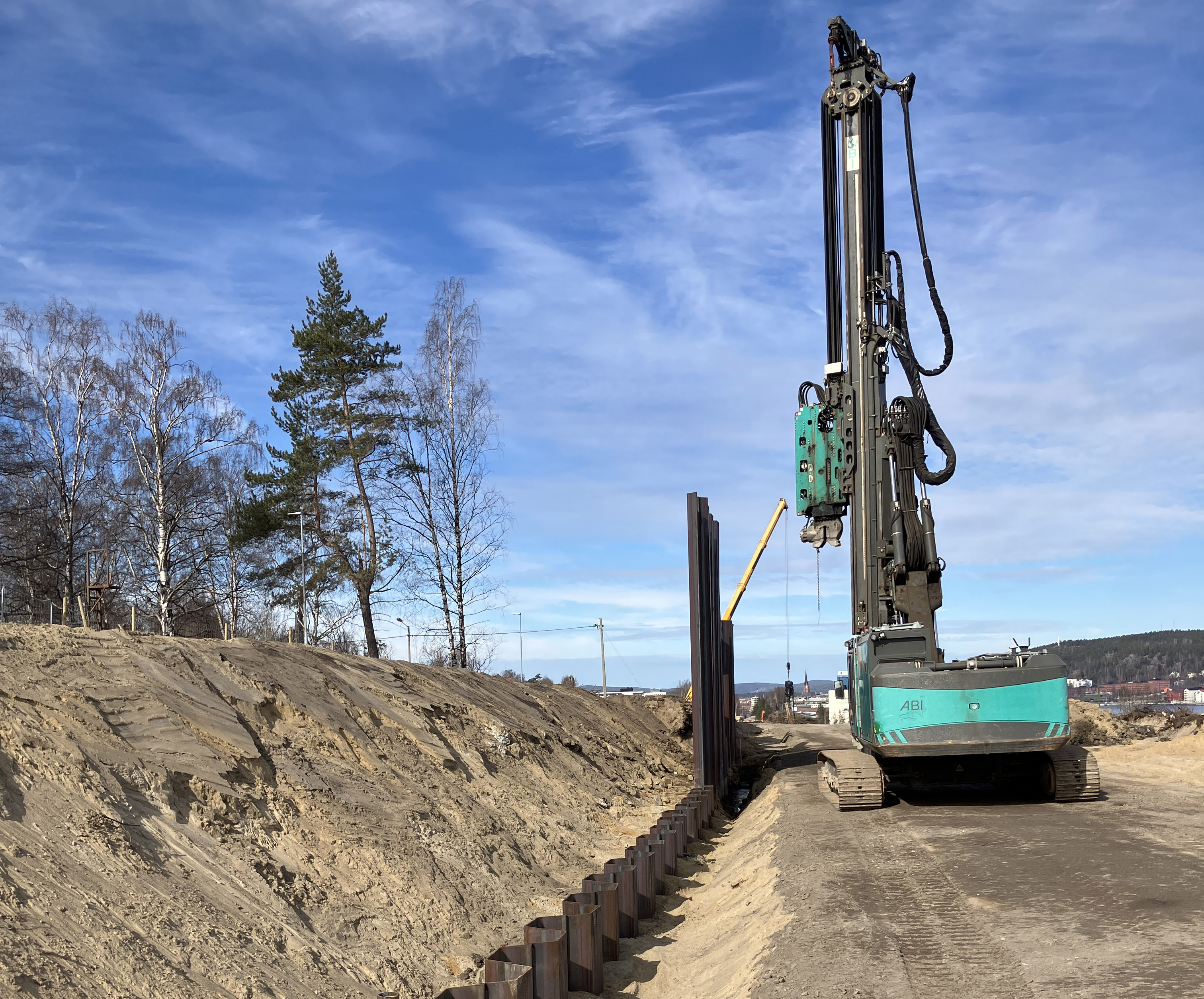 Flera plankor av stål har slagits ner i marken för att bilda en stödmur. Ett par villor nära arbetsområdet som är stängslat. Arbetsmaskiner i bild samt en grupp människor i varselkläder och hjälm på studiebesök. Sundsvallsbron skymtar i bakgrunden.