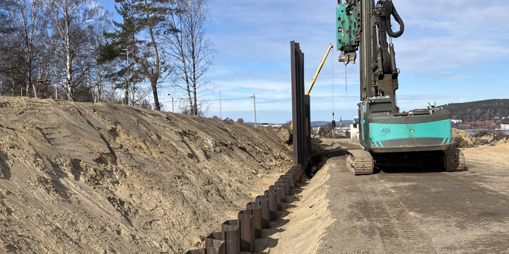 Flera plankor av stål har slagits ner i marken för att bilda en stödmur. Ett par villor nära arbetsområdet som är stängslat. Arbetsmaskiner i bild samt en grupp människor i varselkläder och hjälm på studiebesök. Sundsvallsbron skymtar i bakgrunden.