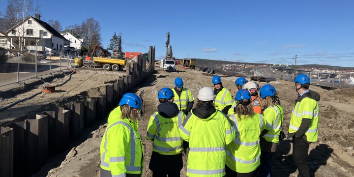 Flera plankor av stål har slagits ner i marken för att bilda en stödmur. Ett par villor nära arbetsområdet som är stängslat. Arbetsmaskiner i bild samt en grupp människor i varselkläder och hjälm på studiebesök. Sundsvallsbron skymtar i bakgrunden.