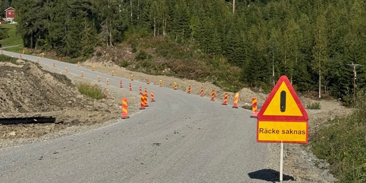 Grusväg som svänger i en kurva. En varningsskylt varnar för att vägräcken saknas. Skog i bakgrunden mot en blå himmel.
