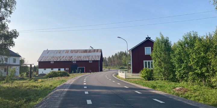 En väg som sträcker sig i en kurva och passerar två hus och en ladugård. Vägen har ett med breda vägrenar.