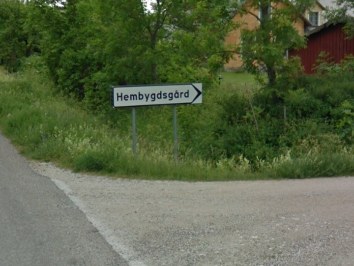 Väg med grönska bakom och en skylt där det står hembygdsgård på.