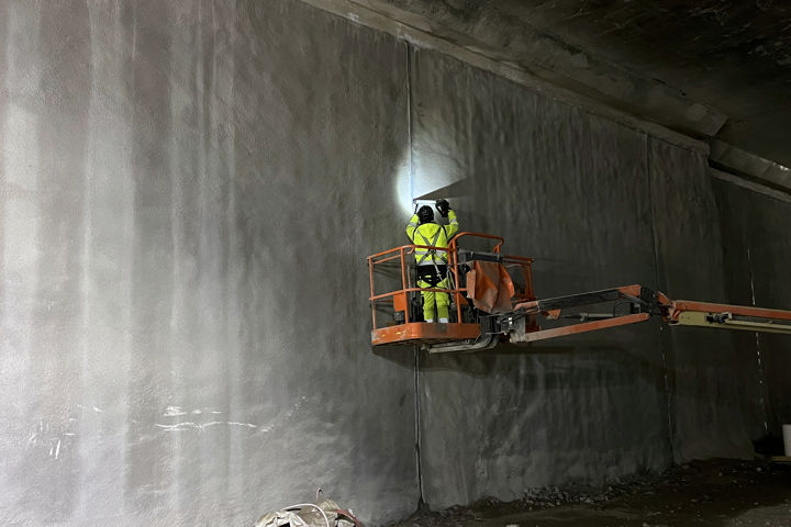 Arbetsklädd person står på en lyftkran och sprutar vätska över betongvägg.